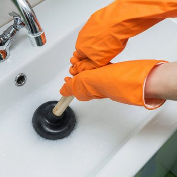 Person wearing orange gloves unblocking a bathroom sink with a plunger. How to Unblock your Bathroom Sink: An Ultimate Tool Guide