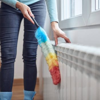 The ultimate guide on how to clean a radiator. Woman cleaning a radiator with a feather duster.