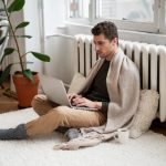 A young man wrapped in a beige blanket sitting cross-legged on a white rug on the floor, typing on a laptop near a radiator next to a potted plant and a warm mug.