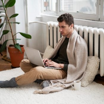 A young man wrapped in a beige blanket sitting cross-legged on a white rug on the floor, typing on a laptop near a radiator next to a potted plant and a warm mug.