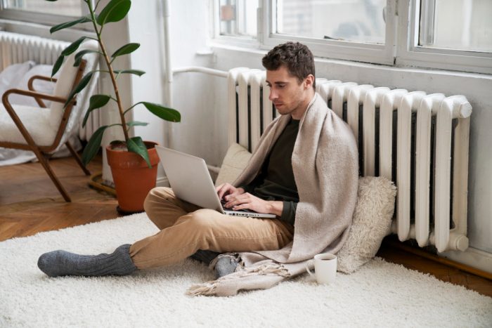 A young man wrapped in a beige blanket sitting cross-legged on a white rug on the floor, typing on a laptop near a radiator next to a potted plant and a warm mug.